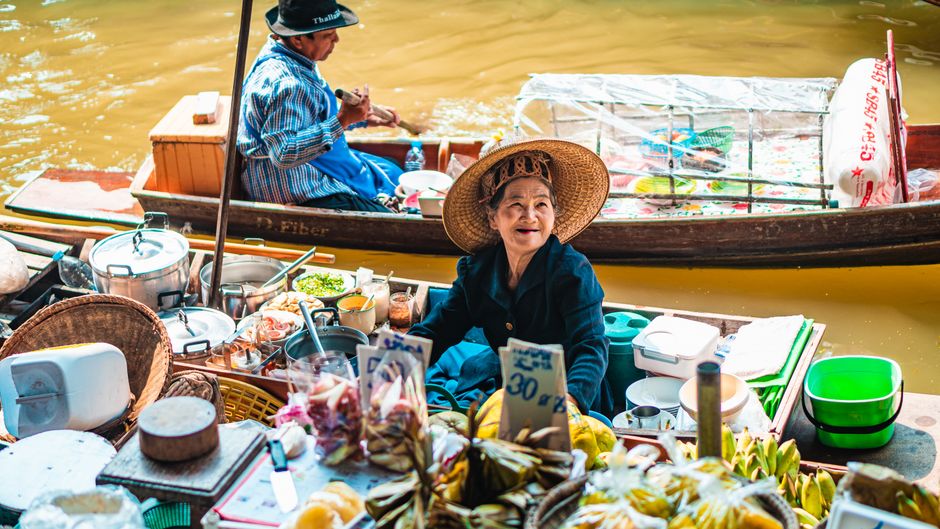 Smiling vendor at Bangkok's floating market selling fresh produce and local dishes from a traditional boat.